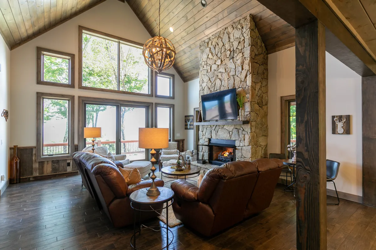 A cozy living room featuring plush leather sofas arranged around a striking stone fireplace. Large windows bathe the space in natural light and offer expansive views of the mountains beyond. Elegant chandeliers and tasteful décor complete the inviting setting.