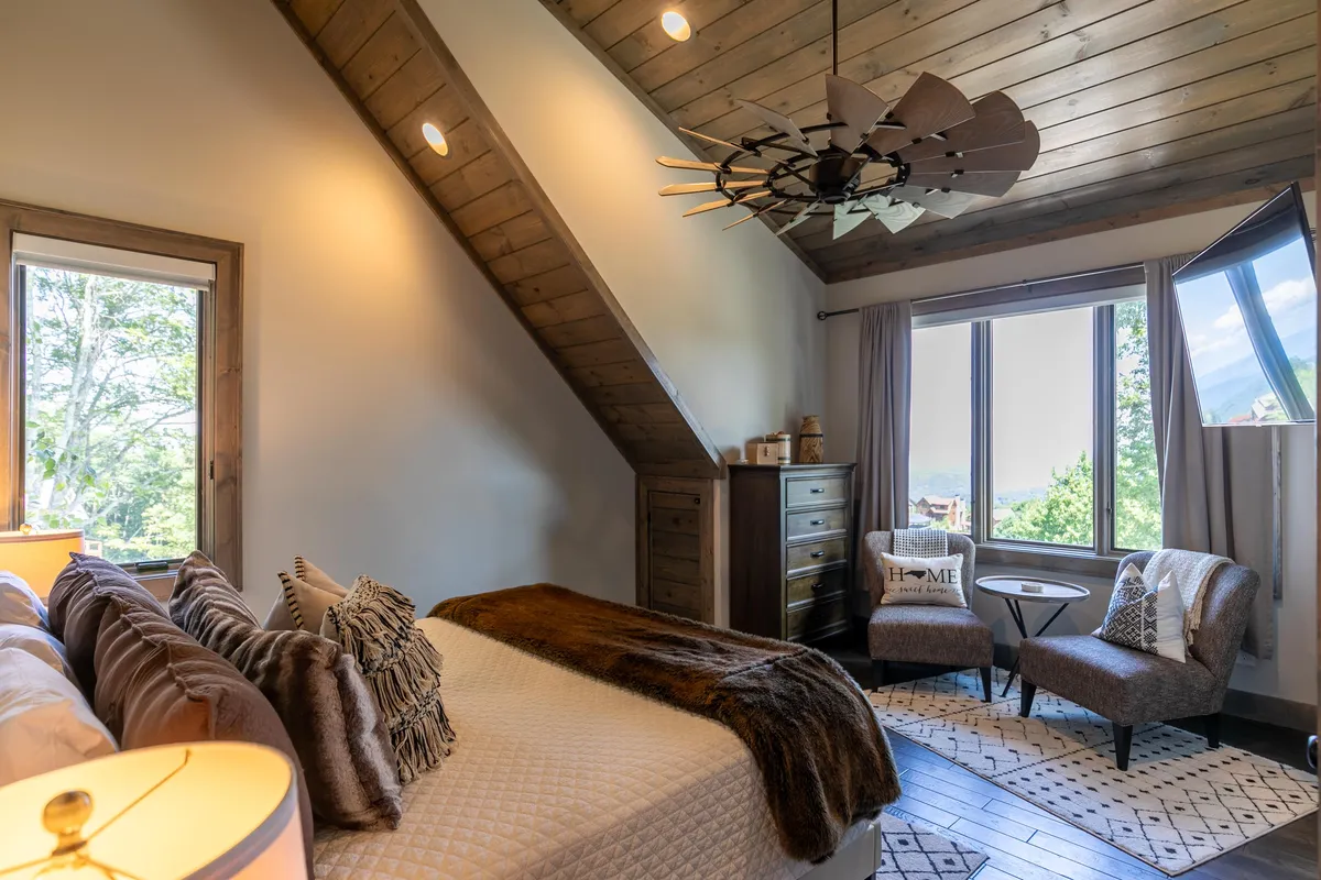 An inviting bedroom with a king bed adorned with decorative pillows, a cozy sitting area with chairs, wooden ceilings, large windows letting in natural light, and a stylish light fixture. The space exudes comfort and serenity, perfect for quiet downtime.