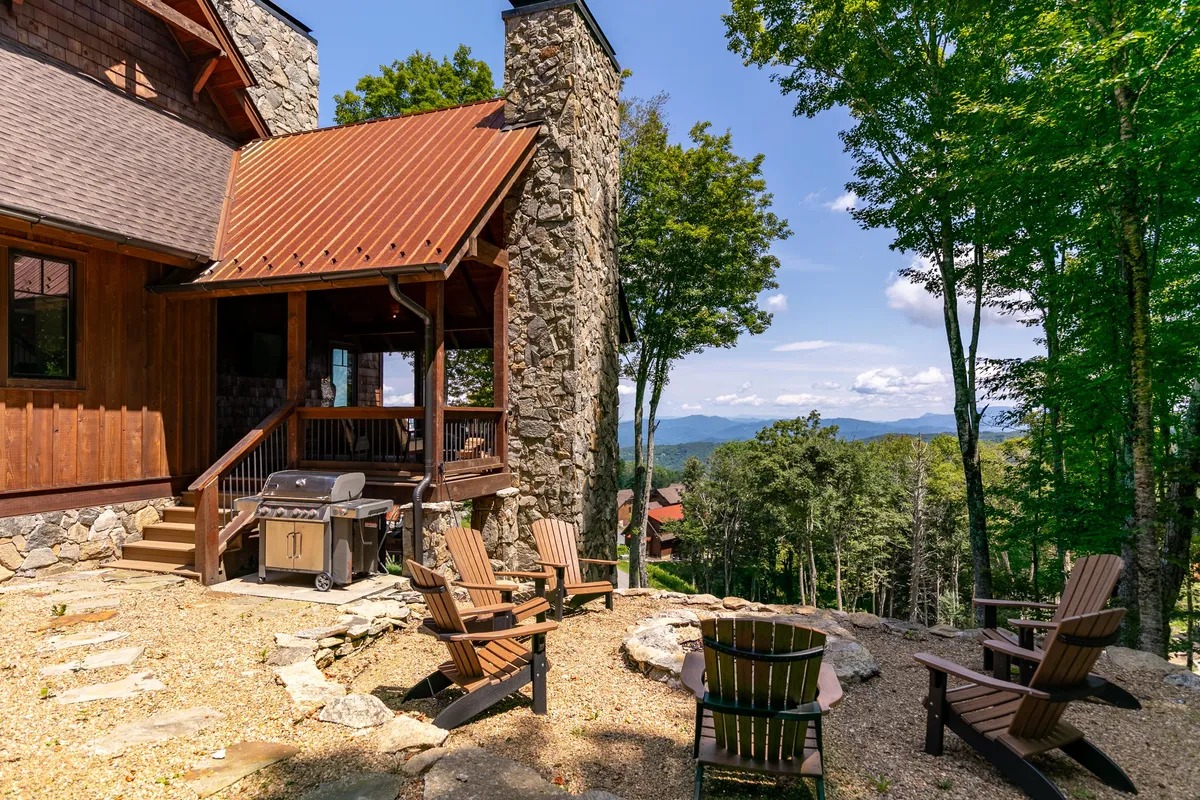 A welcoming wooden deck with a stone chimney, gas grill, fire pit, and Adirondack chairs arrayed for social gatherings. The panoramic backdrop of the mountains enhances the inviting ambiance, making it ideal for cookouts and evening conversations by the fire.