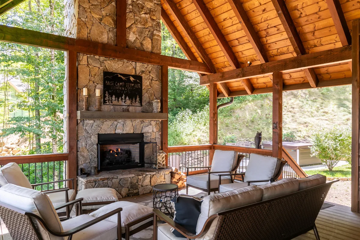 A charming covered porch with a stone fireplace, comfortable seating, and a wooden ceiling, set against a backdrop of greenery. The scene suggests warmth and year-round usability, perfect for morning coffees or evenings by the flickering fire—rain or shine.