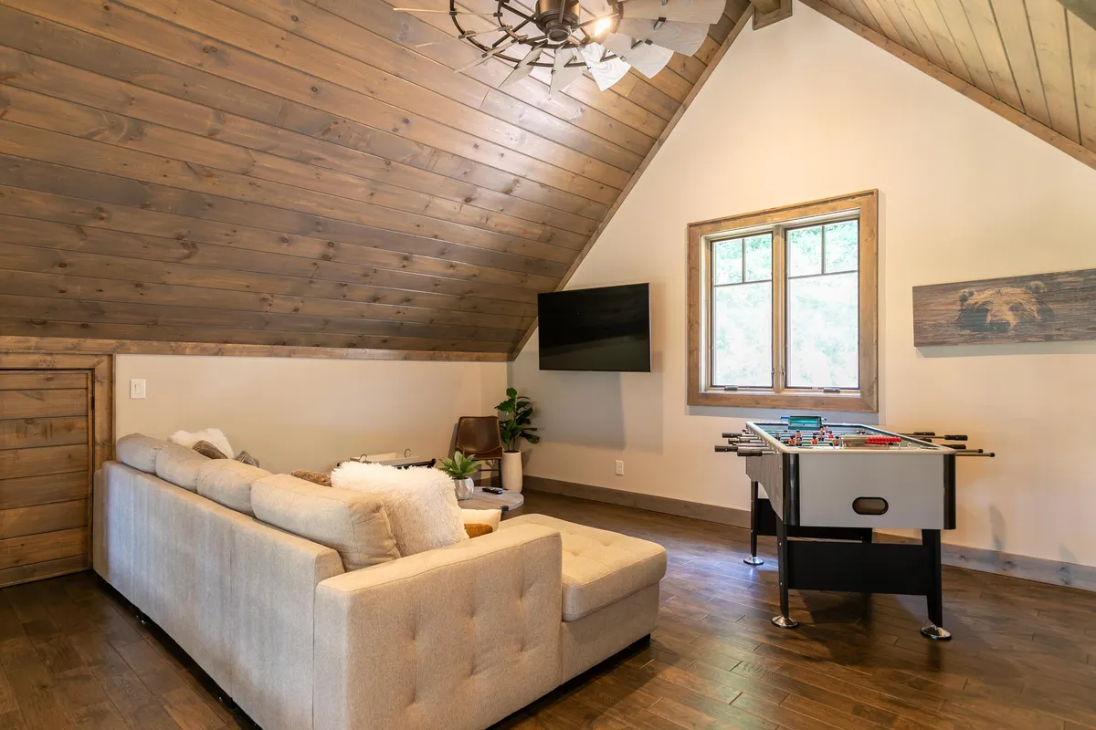 An entertainment loft with a foosball table, comfortable sofa, window for natural light, and a ceiling fan. The room has a relaxed, playful vibe, inviting guests for friendly competition, games, or relaxed lounging with a movie.