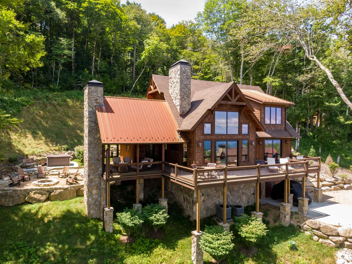 The image showcases a stunning cabin set amidst lush trees, featuring expansive windows, a striking stone chimney, and a spacious wooden deck. The outdoor living area includes a cozy fire pit with inviting seating, making it perfect for gathering under the stars. The natural setting hints at beautiful sunset views and serene mountain tranquility.