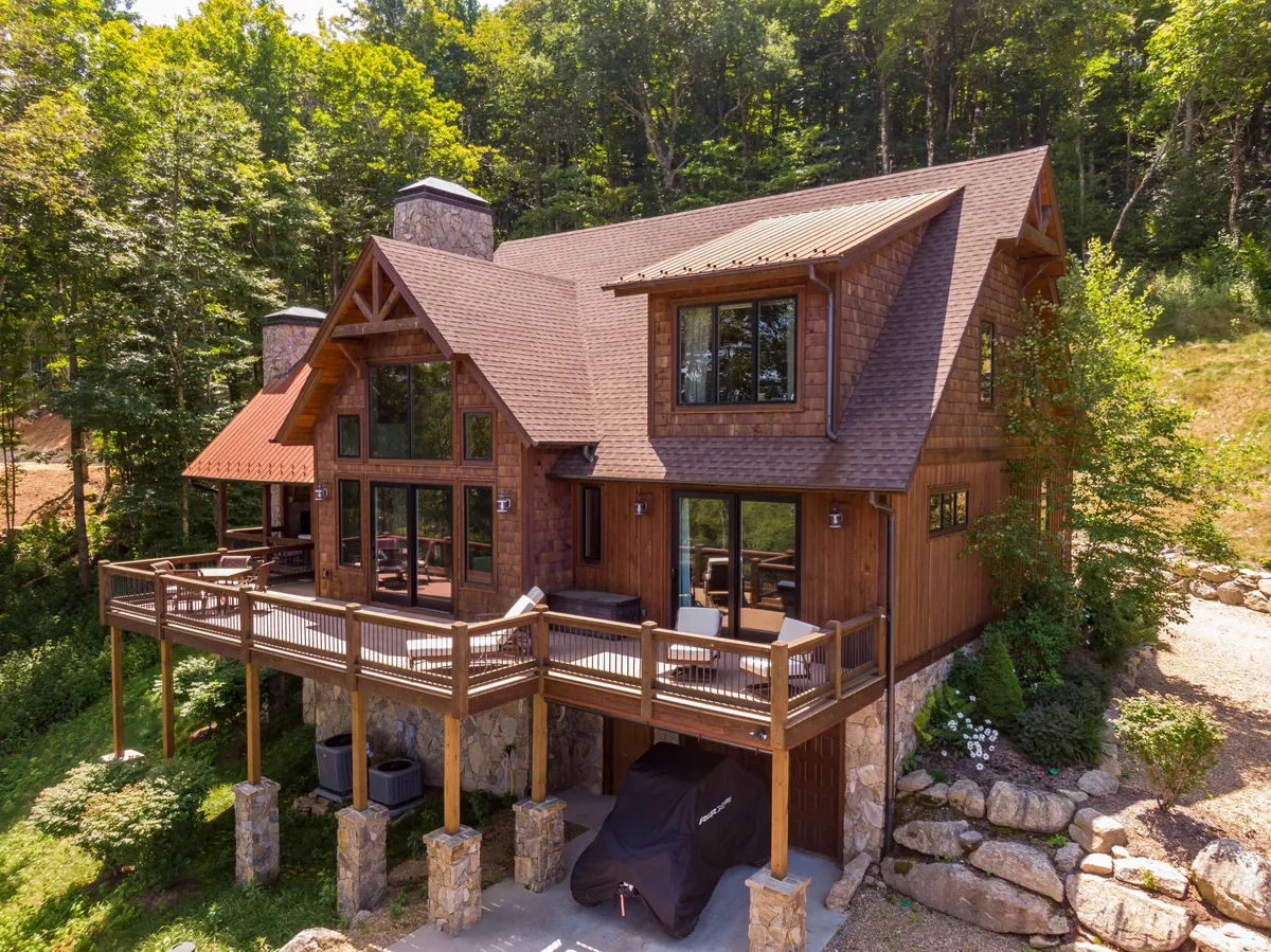 This image features the expansive front deck of the property, showcasing its sturdy stone foundation, wooden siding, and large windows. The outdoor scene exudes a relaxed, welcoming atmosphere, with areas for lounging and barbecuing while taking in the natural mountain surroundings.