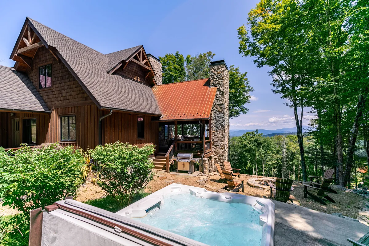 A spacious wooden deck featuring a bubbling hot tub, a stone chimney, and sweeping views of lush mountains and greenery. Outdoor dining furniture and a fire pit complete the cozy, inviting setup. The scene is tranquil, perfect for relaxing after a day of mountain adventures.