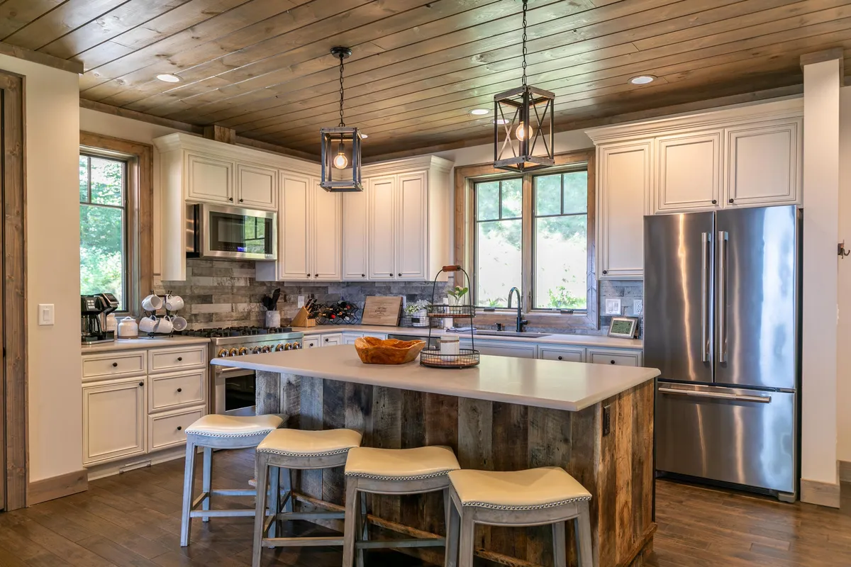 A spacious, sunlit kitchen with a prominent granite island at its center. Wooden cabinets and stylish hanging light fixtures enhance the warm, inviting atmosphere. Large windows frame striking mountain views, and the open layout connects seamlessly to adjacent living and dining areas.