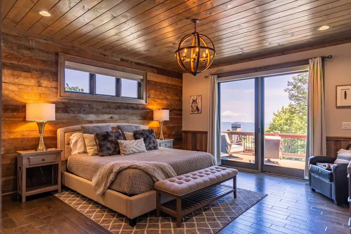 A serene bedroom retreat featuring a plush bed, soft bedding, large windows framing a scenic view, a stylish chandelier above, and a rustic wooden wall. The morning light pours through the windows, creating a tranquil atmosphere ideal for restful sleep and peaceful mornings.