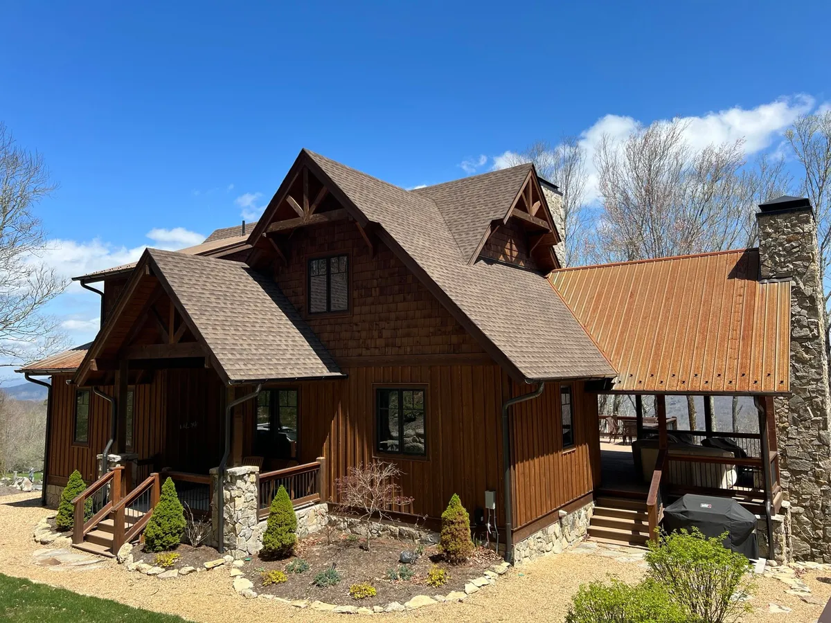 The exterior of a charming wooden home is accented with stone details and nestled within lush greenery. A spacious porch with comfortable seating invites groups to gather, enjoy mountain views, or stargaze in a cozy, communal setting.