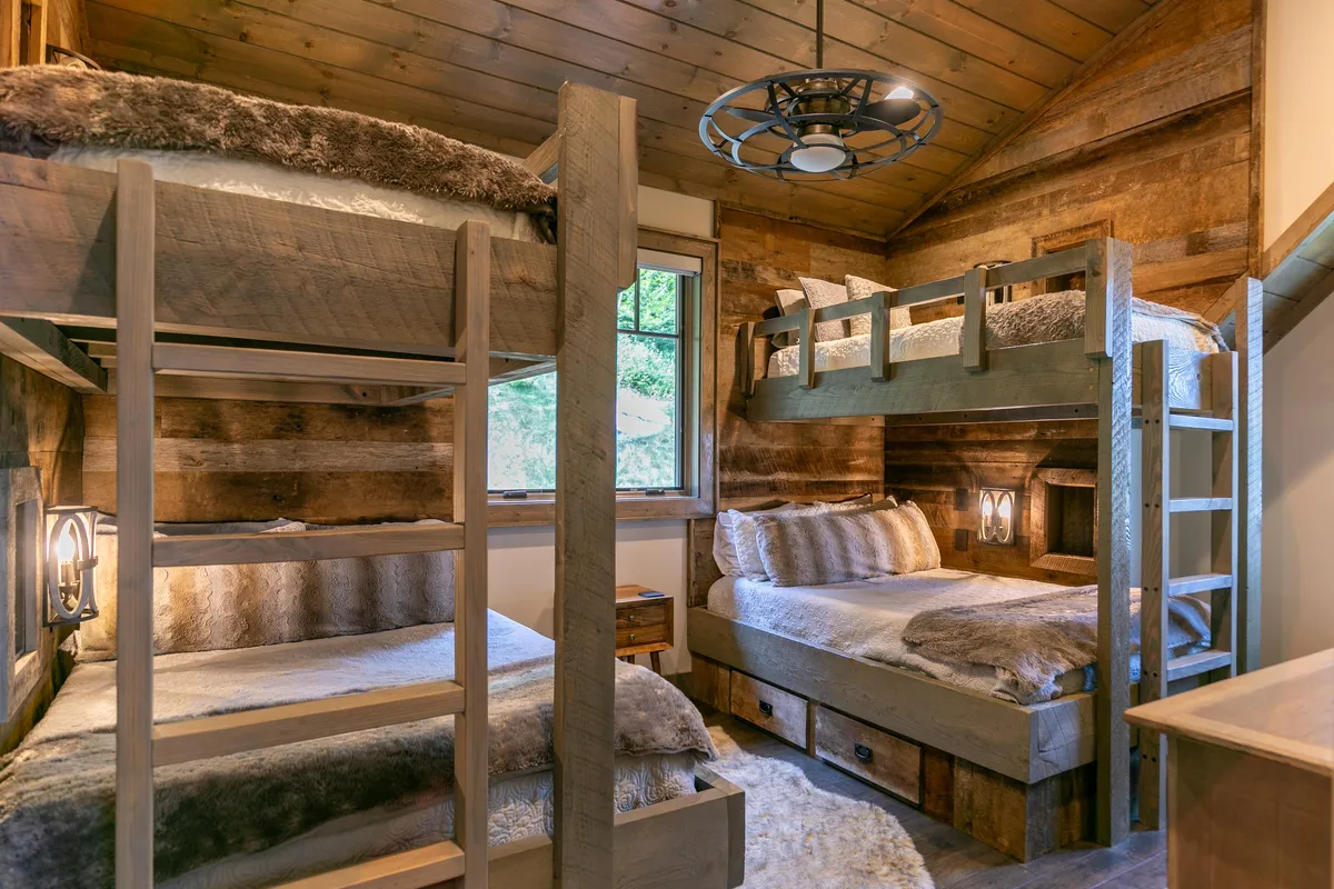 A warm and inviting bunk room with sturdy wooden bunk beds, sunlight streaming in through a window, and natural wood textures throughout. The room is designed for comfort and group fun, suitable for children or groups.