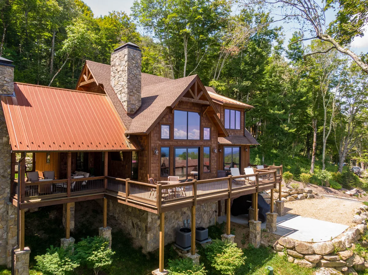 This exterior image captures the rustic wooden architecture of the home, large windows, a broad wooden deck, and a stone chimney. The deck is set up for outdoor dining and relaxation, with mountain scenery in the background and hints of nearby hiking trails.