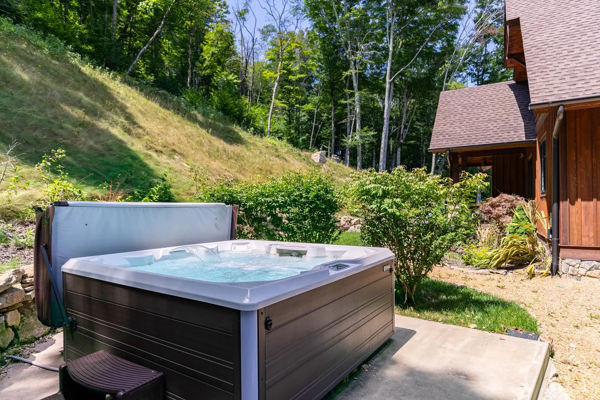 A bubbling hot tub sits on a wooden deck, surrounded by vibrant green trees and the exterior of a cozy wooden house. The inviting setting encourages guests to unwind together under the open sky, whether for early morning relaxation or evening stargazing sessions.